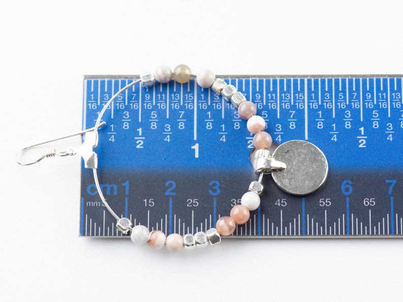 Pu&ograve; includere: Un orecchino a cerchio d'argento con un ciondolo e una fila di perline rosa e bianche. L'orecchino &egrave; misurato con un righello, mostrando che ha un diametro di 3 cm.