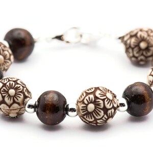 Op de afbeelding: Een bruin en witte kralen armband met bloemmotieven. De armband heeft een zilveren sluiting.