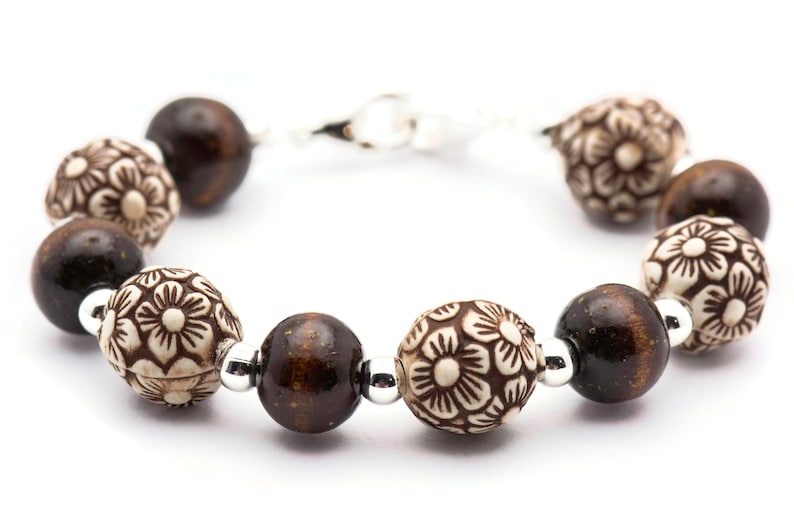 Op de afbeelding: Een bruin en witte kralen armband met bloemmotieven. De armband heeft een zilveren sluiting.