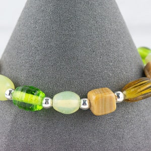 May include: A green, brown and white beaded bracelet with silver spacers. The bracelet is displayed on a grey cone.