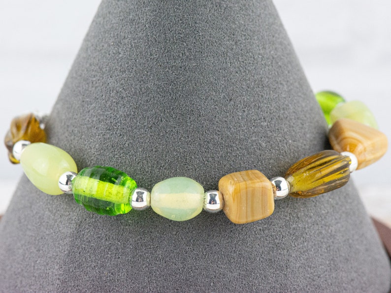 May include: A green, brown and white beaded bracelet with silver spacers. The bracelet is displayed on a grey cone.