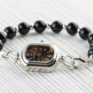 May include: A silver watch with a rectangular face and a brown dial. The watch is attached to a bracelet of large, round, black beads. The watch face reads "Geneva".