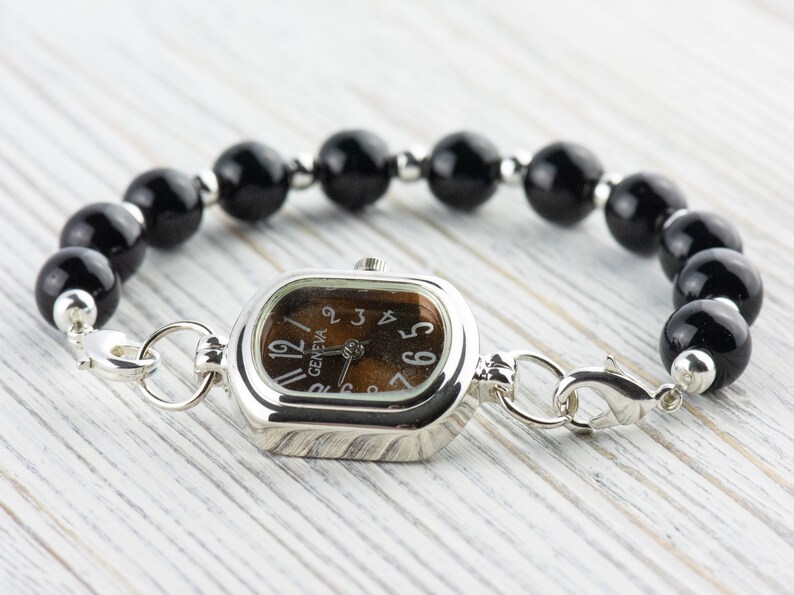 May include: A silver watch with a rectangular face and a brown dial. The watch is attached to a bracelet of large, round, black beads. The watch face reads "Geneva".