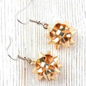 May include: A pair of gold-colored bow earrings with silver-colored ear wires. The earrings are made of a shiny, metallic material and are designed to resemble gift bows. The earrings are displayed on a white, textured surface.