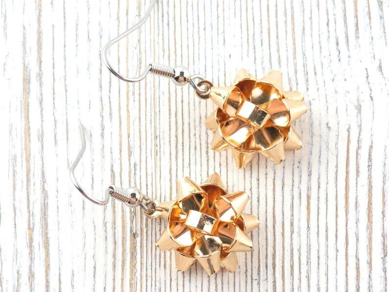 May include: A pair of gold-colored bow earrings with silver-colored ear wires. The earrings are made of a shiny, metallic material and are designed to resemble gift bows. The earrings are displayed on a white, textured surface.