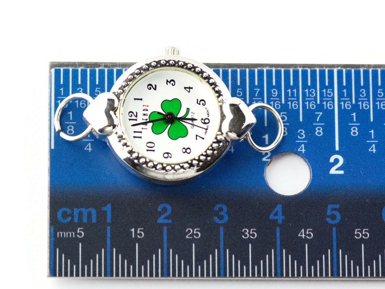 Peut inclure: Une montre argent&eacute;e avec un cadran blanc et un tr&egrave;fle vert. La montre mesure 2,5 cm de diam&egrave;tre et a un fermoir en forme de c&oelig;ur.