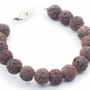Puede incluir: Una pulsera con cuentas de roca volc&aacute;nica marr&oacute;n oscuro y porosa. Las cuentas son redondas y tienen una superficie texturizada. La pulsera est&aacute; ensartada en una cadena plateada y tiene un cierre de broche.