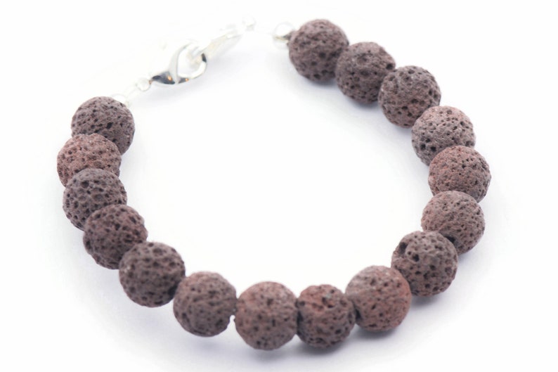 Puede incluir: Una pulsera con cuentas de roca volc&aacute;nica marr&oacute;n oscuro y porosa. Las cuentas son redondas y tienen una superficie texturizada. La pulsera est&aacute; ensartada en una cadena plateada y tiene un cierre de broche.
