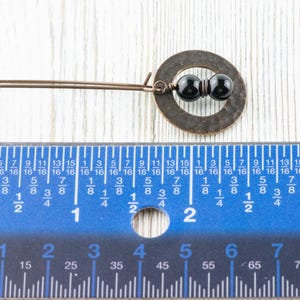 Peut inclure: Boucle d'oreille de couleur cuivre faite &agrave; la main, avec un cercle et deux perles noires. La boucle d'oreille mesure environ 5 cm de long, comme indiqu&eacute; par la r&egrave;gle. Pos&eacute;e sur une surface en bois blanc.