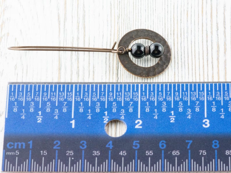 Peut inclure: Boucle d'oreille de couleur cuivre faite &agrave; la main, avec un cercle et deux perles noires. La boucle d'oreille mesure environ 5 cm de long, comme indiqu&eacute; par la r&egrave;gle. Pos&eacute;e sur une surface en bois blanc.