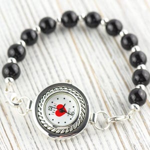 May include: A quartz watch with a black and silver bezel and a white face featuring a red heart. The watch band is made of black beads and silver links. The watch is displayed on a light wood surface.