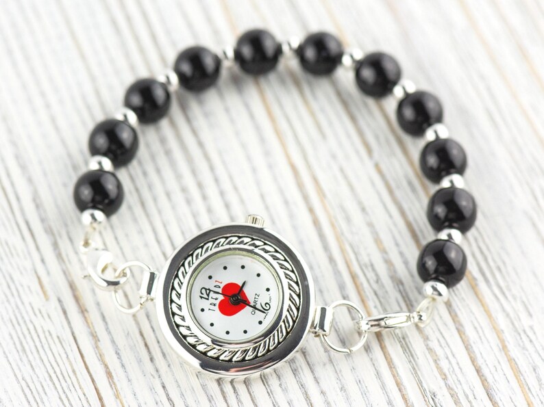 May include: A quartz watch with a black and silver bezel and a white face featuring a red heart. The watch band is made of black beads and silver links. The watch is displayed on a light wood surface.