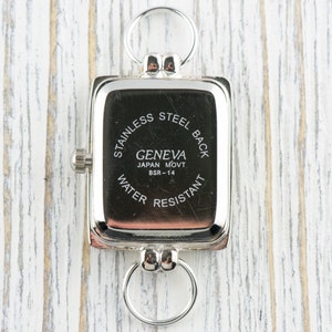 May include: A rectangular silver-toned watch with a stainless steel back and a ring at the top and bottom. The watch face is black with white text that reads "GENEVA JAPAN MOVT BSR-14 WATER RESISTANT."