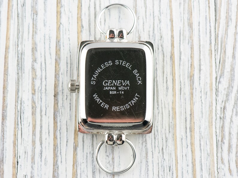 May include: A rectangular silver-toned watch with a stainless steel back and a ring at the top and bottom. The watch face is black with white text that reads "GENEVA JAPAN MOVT BSR-14 WATER RESISTANT."