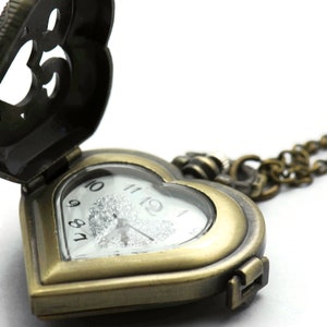 Puede incluir: Un reloj de bolsillo antiguo en forma de coraz&oacute;n con la tapa abierta. El reloj tiene un acabado en bronce y una esfera blanca con n&uacute;meros negros. Una cadena de bronce est&aacute; unida al reloj.