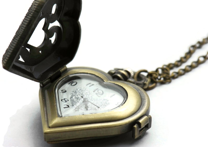 Puede incluir: Un reloj de bolsillo antiguo en forma de coraz&oacute;n con la tapa abierta. El reloj tiene un acabado en bronce y una esfera blanca con n&uacute;meros negros. Una cadena de bronce est&aacute; unida al reloj.