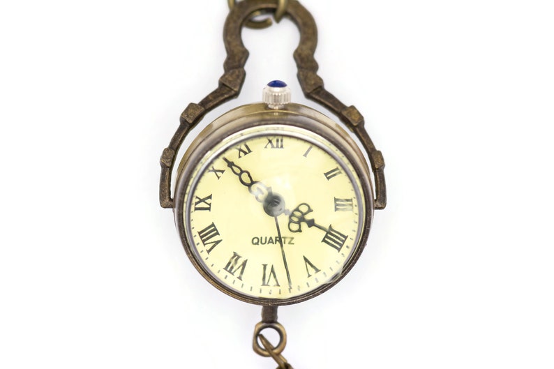 May include: A gold-toned pocket watch with Roman numeral markings on the face. The watch face is cream-coloured with black hands and the word "QUARTZ" printed in black.