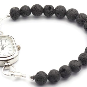 May include: A silver watch with a square face and black Roman numeral markings. The watch is attached to a black lava rock beaded bracelet.