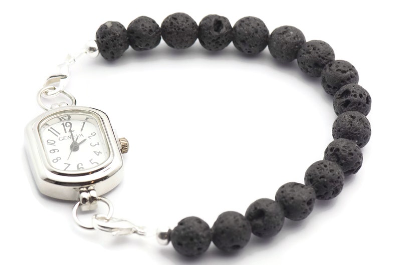 May include: A silver watch with a square face and black Roman numeral markings. The watch is attached to a black lava rock beaded bracelet.