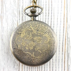 May include: Antique-style pocket watch with an ornate floral design on the cover. The watch has a bronze finish and hangs from a matching chain. The watch face is not visible in this image.