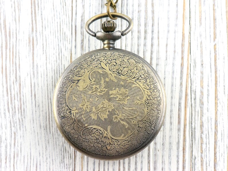 May include: Antique-style pocket watch with an ornate floral design on the cover. The watch has a bronze finish and hangs from a matching chain. The watch face is not visible in this image.