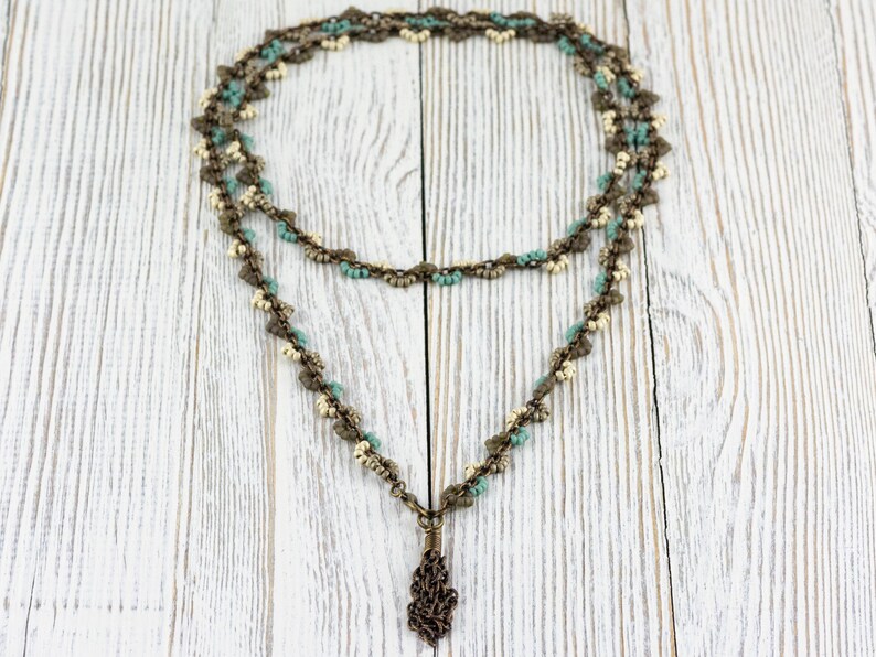 May include: A double-strand necklace with a pendant. The necklace is made of small, light blue and white beads, and the pendant is a bronze-coloured, intricate design.