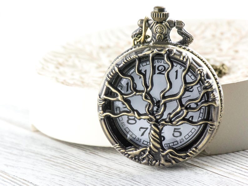 May include: A bronze pocket watch featuring a tree of life design. The watch face is white with black numerals and hands. The watch is set on a white surface. The watch's diameter is approximately 4 cm.