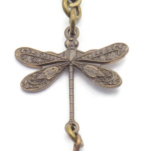 Puede incluir: Un amuleto de lib&eacute;lula de bronce con una cuenta de piedra rosa y marr&oacute;n que cuelga. El amuleto est&aacute; unido a un gancho de bronce.