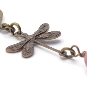Puede incluir: Un par de pendientes colgantes con un dije de lib&eacute;lula y una cuenta de piedra rosa y marr&oacute;n. La lib&eacute;lula est&aacute; hecha de lat&oacute;n y tiene un dise&ntilde;o detallado. Los pendientes est&aacute;n unidos a un gancho de plata.