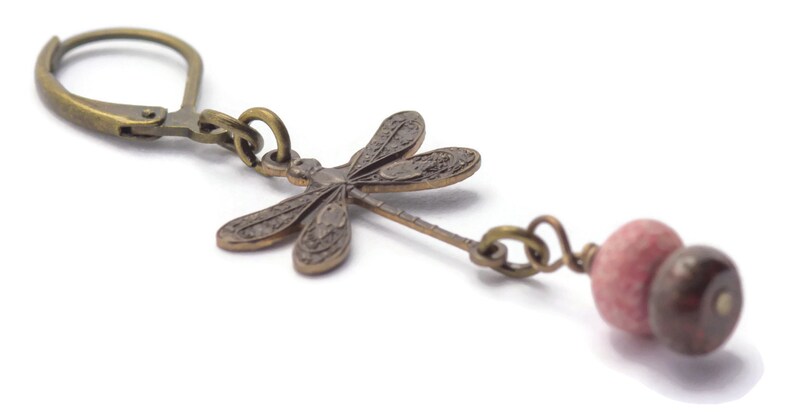 Puede incluir: Un par de pendientes colgantes con un dije de lib&eacute;lula y una cuenta de piedra rosa y marr&oacute;n. La lib&eacute;lula est&aacute; hecha de lat&oacute;n y tiene un dise&ntilde;o detallado. Los pendientes est&aacute;n unidos a un gancho de plata.