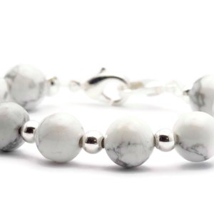 May include: A bracelet featuring white and grey marbled stone beads, interspersed with silver-coloured metal beads and a silver clasp. The beads are round. A piece of jewellery.
