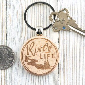 May include: A wooden keyring with a round, light brown tag that says "River Life" in cursive script. The tag also features a laser-engraved image of a river winding through a forest. The keyring is attached to a silver key ring.