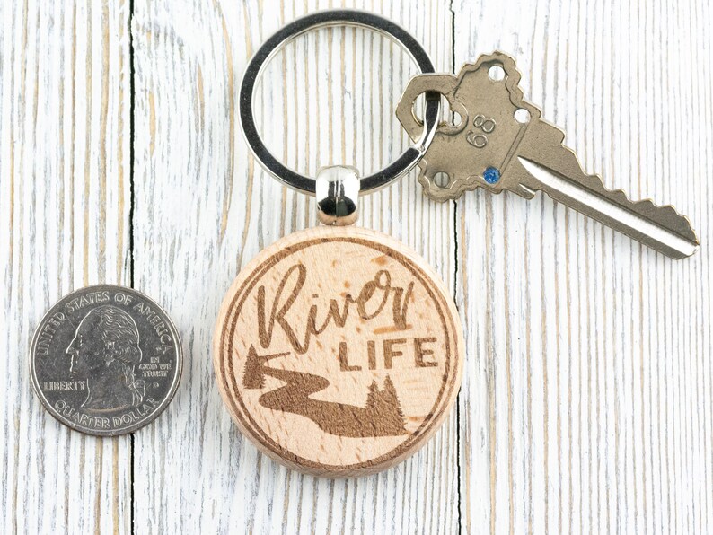 May include: A wooden keyring with a round, light brown tag that says "River Life" in cursive script. The tag also features a laser-engraved image of a river winding through a forest. The keyring is attached to a silver key ring.