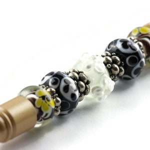 May include: A gold pen with black, white, and yellow glass beads. The beads have different patterns and are separated by silver spacers.