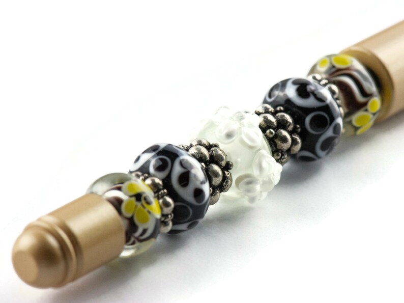 May include: A gold pen with black, white, and yellow glass beads. The beads have different patterns and are separated by silver spacers.