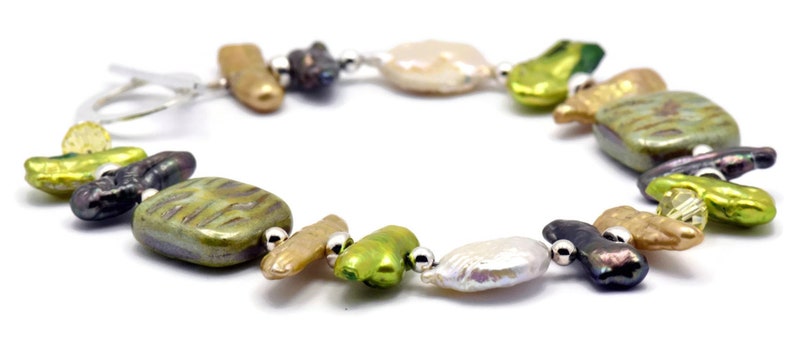 K&ouml;nnte beinhalten: Ein Armband aus gr&uuml;nen, braunen und wei&szlig;en S&uuml;&szlig;wasserperlen mit silbernen Akzenten. Das Armband verf&uuml;gt &uuml;ber eine Vielzahl von Formen und Gr&ouml;&szlig;en von Perlen, darunter runde, ovale und quadratische.