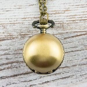 May include: A gold-toned pocket watch with a chain hanging from it. The watch is on a white wooden background.