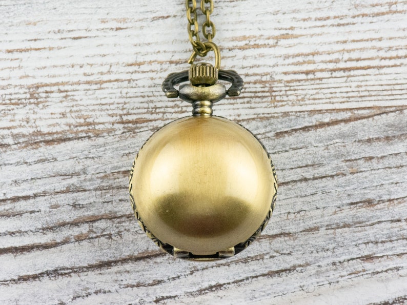 May include: A gold-toned pocket watch with a chain hanging from it. The watch is on a white wooden background.