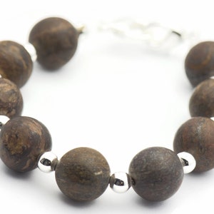 Puede incluir: Un brazalete de cuentas de madera marr&oacute;n con detalles plateados. El brazalete est&aacute; hecho de cuentas redondas con un patr&oacute;n de veta de madera natural.