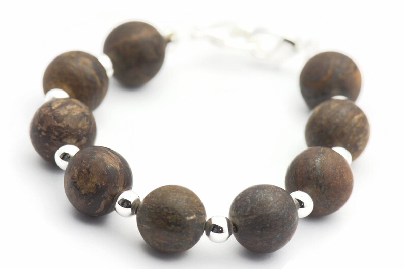 Puede incluir: Un brazalete de cuentas de madera marr&oacute;n con detalles plateados. El brazalete est&aacute; hecho de cuentas redondas con un patr&oacute;n de veta de madera natural.