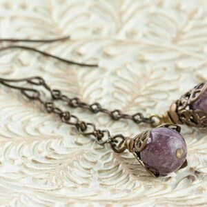 May include: Handmade earrings with small, round, purple gemstones. The gemstones are set in decorative, filigree-style metalwork, hanging from fine chain links and ear wires. The metal has a warm, antique bronze finish.