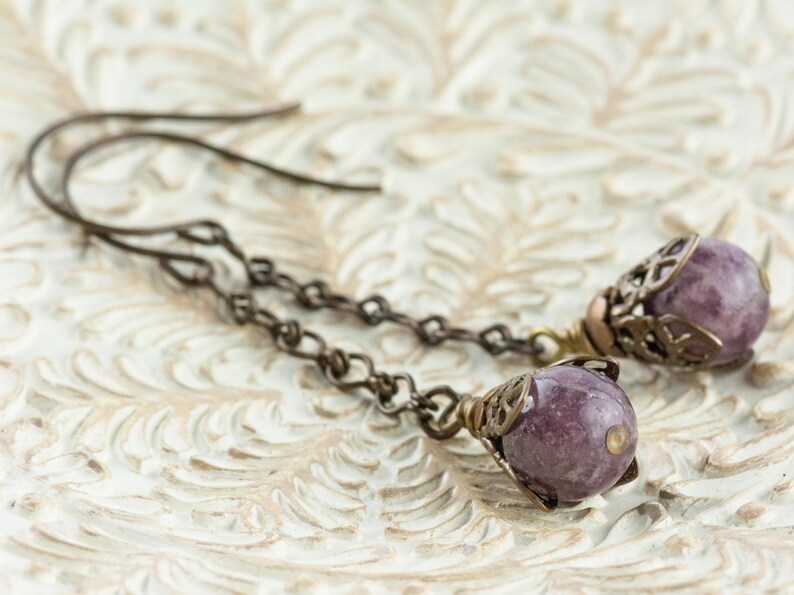 May include: Handmade earrings with small, round, purple gemstones. The gemstones are set in decorative, filigree-style metalwork, hanging from fine chain links and ear wires. The metal has a warm, antique bronze finish.