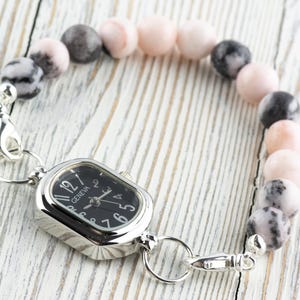 May include: A beaded bracelet watch with a black watch face and silver-tone metal accents. The bracelet features a mix of pink, gray, and black round beads. The watch face has the word "Geneva" printed on it.