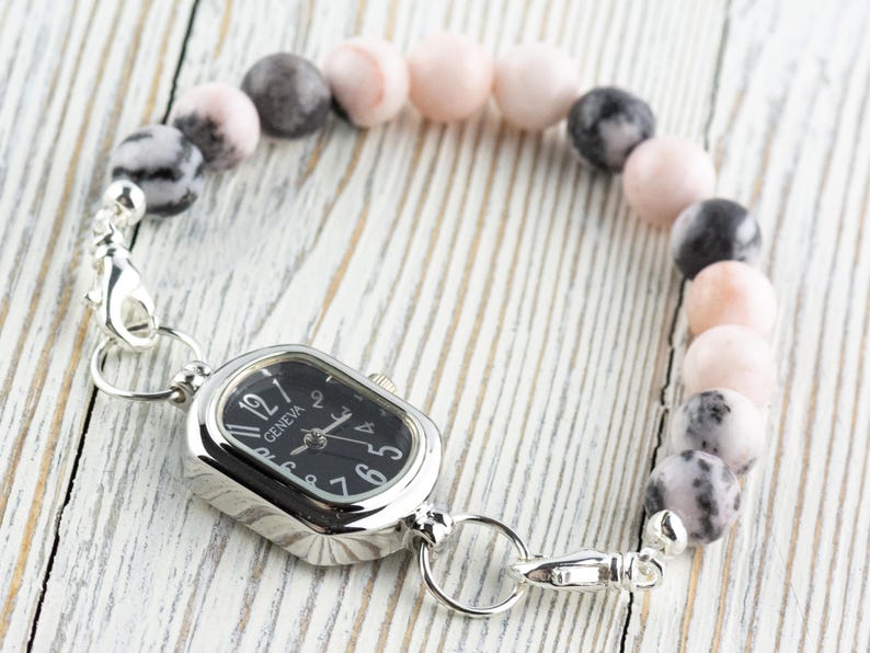 May include: A beaded bracelet watch with a black watch face and silver-tone metal accents. The bracelet features a mix of pink, gray, and black round beads. The watch face has the word "Geneva" printed on it.