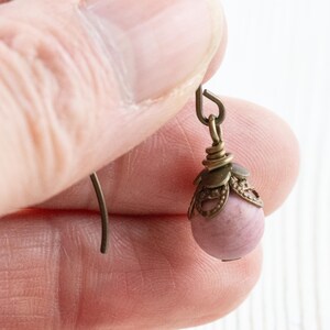 K&ouml;nnte beinhalten: Nahaufnahme eines einzelnen Ohrrings. Der Ohrring hat eine kleine, runde, hellrosa Perle, die an einer dekorativen, bronzefarbenen Metallkappe und einem Haken h&auml;ngt. Das Metall hat ein Vintage-Aussehen. Der Ohrring wird von einer Person gehalten.
