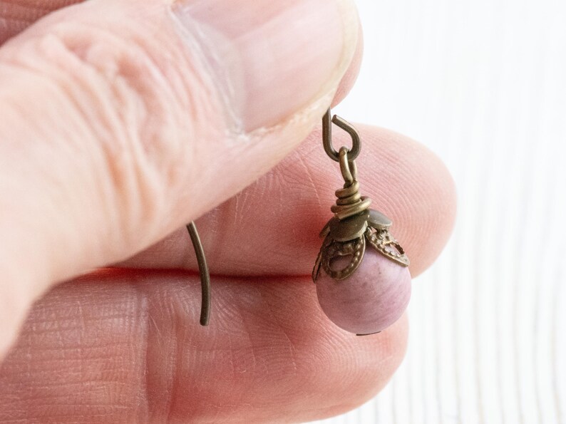 K&ouml;nnte beinhalten: Nahaufnahme eines einzelnen Ohrrings. Der Ohrring hat eine kleine, runde, hellrosa Perle, die an einer dekorativen, bronzefarbenen Metallkappe und einem Haken h&auml;ngt. Das Metall hat ein Vintage-Aussehen. Der Ohrring wird von einer Person gehalten.