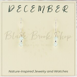 May include: A pair of silver dangle earrings with two light blue crystal beads on each earring. The earrings are displayed on a card that says "December" and "Black Brook Shop Handmade Jewelry".