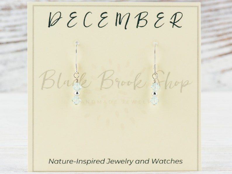 May include: A pair of silver dangle earrings with two light blue crystal beads on each earring. The earrings are displayed on a card that says "December" and "Black Brook Shop Handmade Jewelry".
