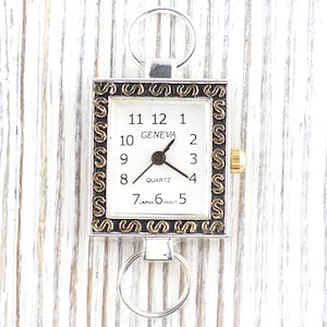 Peut inclure: Une montre carr&eacute;e Geneva &agrave; quartz avec un cadran blanc, des chiffres noirs et des accents dor&eacute;s. La montre a un cadre argent&eacute; avec des d&eacute;tails d&eacute;coratifs et un anneau argent&eacute; en haut et en bas. La montre indique "GENEVA" et "JAPAN MOVT".