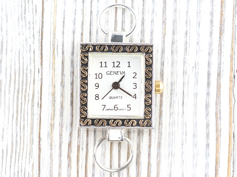 Peut inclure: Une montre carr&eacute;e Geneva &agrave; quartz avec un cadran blanc, des chiffres noirs et des accents dor&eacute;s. La montre a un cadre argent&eacute; avec des d&eacute;tails d&eacute;coratifs et un anneau argent&eacute; en haut et en bas. La montre indique "GENEVA" et "JAPAN MOVT".